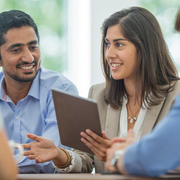 A multi-ethnic group of young business men and women in semi-casual office clothes are sharing ideas and holding a digital tablet in an indoor, sunlit office.